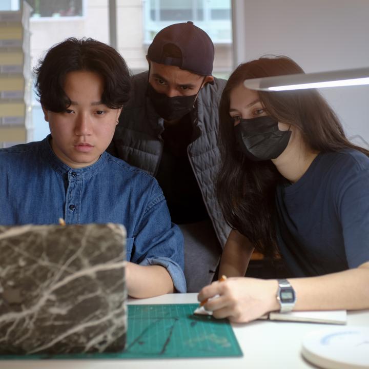 Three graduate students, masked and unmasked, looking at laptop screen