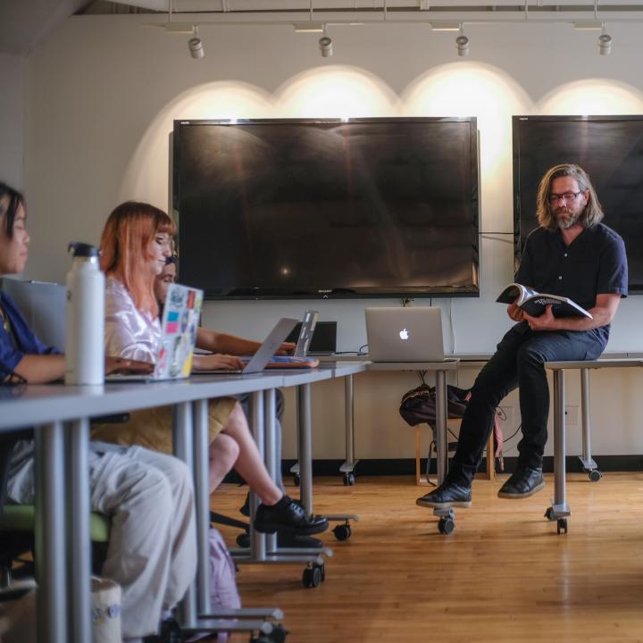 Graduate class, students on laptops while instructors sit on table. 