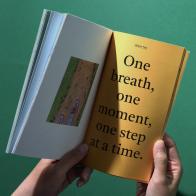 Hands holding up an orange book. The words on the page read: One breath, one moment, one step at a time.